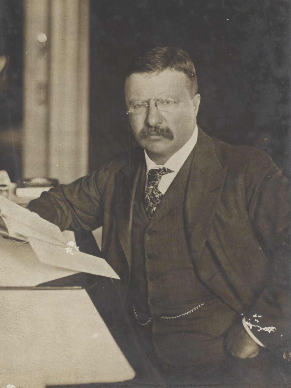 President Roosevelt holds papers while seated at his desk.